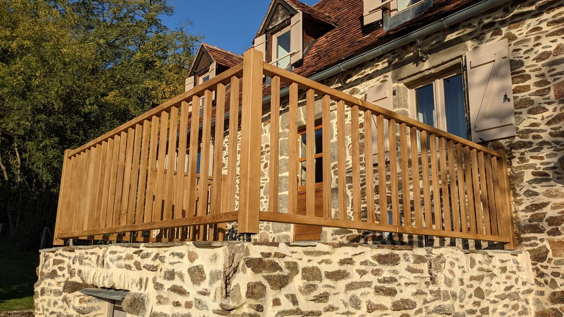 Maison en pierre avec une grande terrasse en bois et une balustrade donnant sur une cour ensoleillée