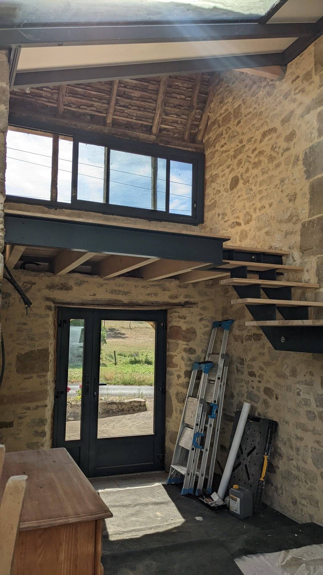 Intérieur de loft aux murs en pierre avec escalier, poutres apparentes et hautes fenêtres au-dessus d'une porte vitrée