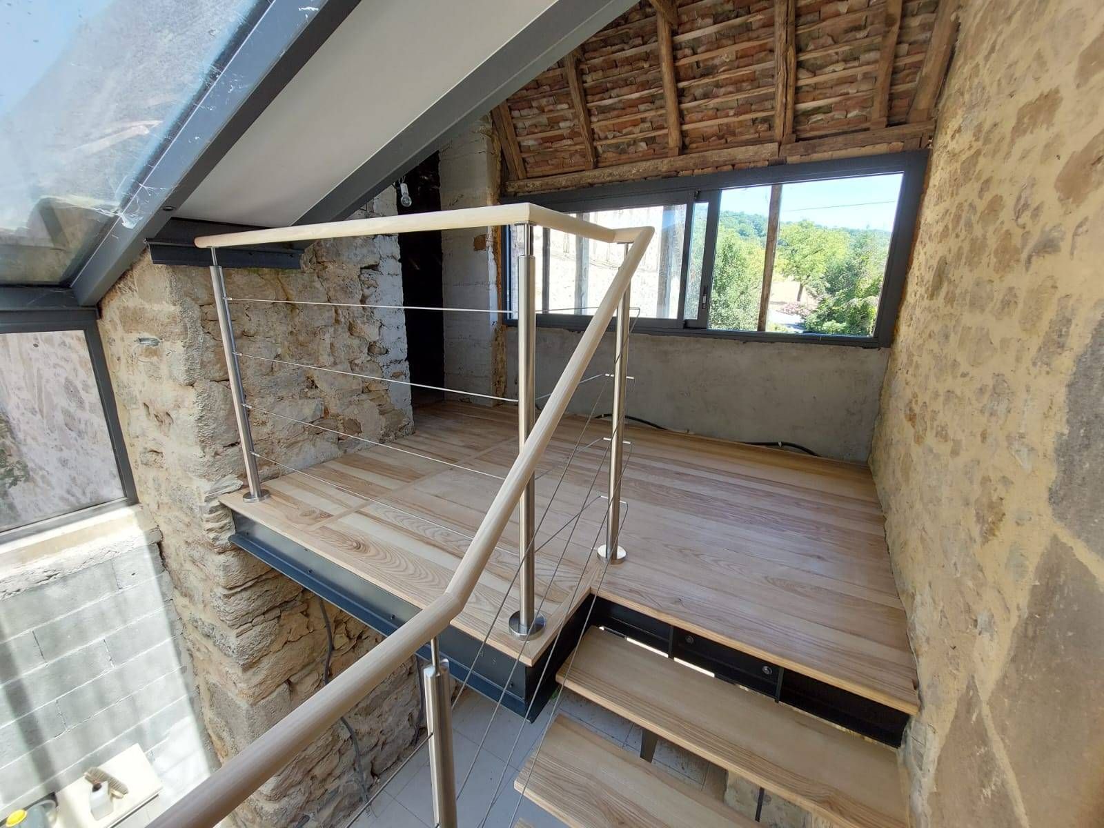 Intérieur d'un loft aux murs en pierre, avec estrade en bois, rambarde et grande fenêtre sous un plafond mansardé.