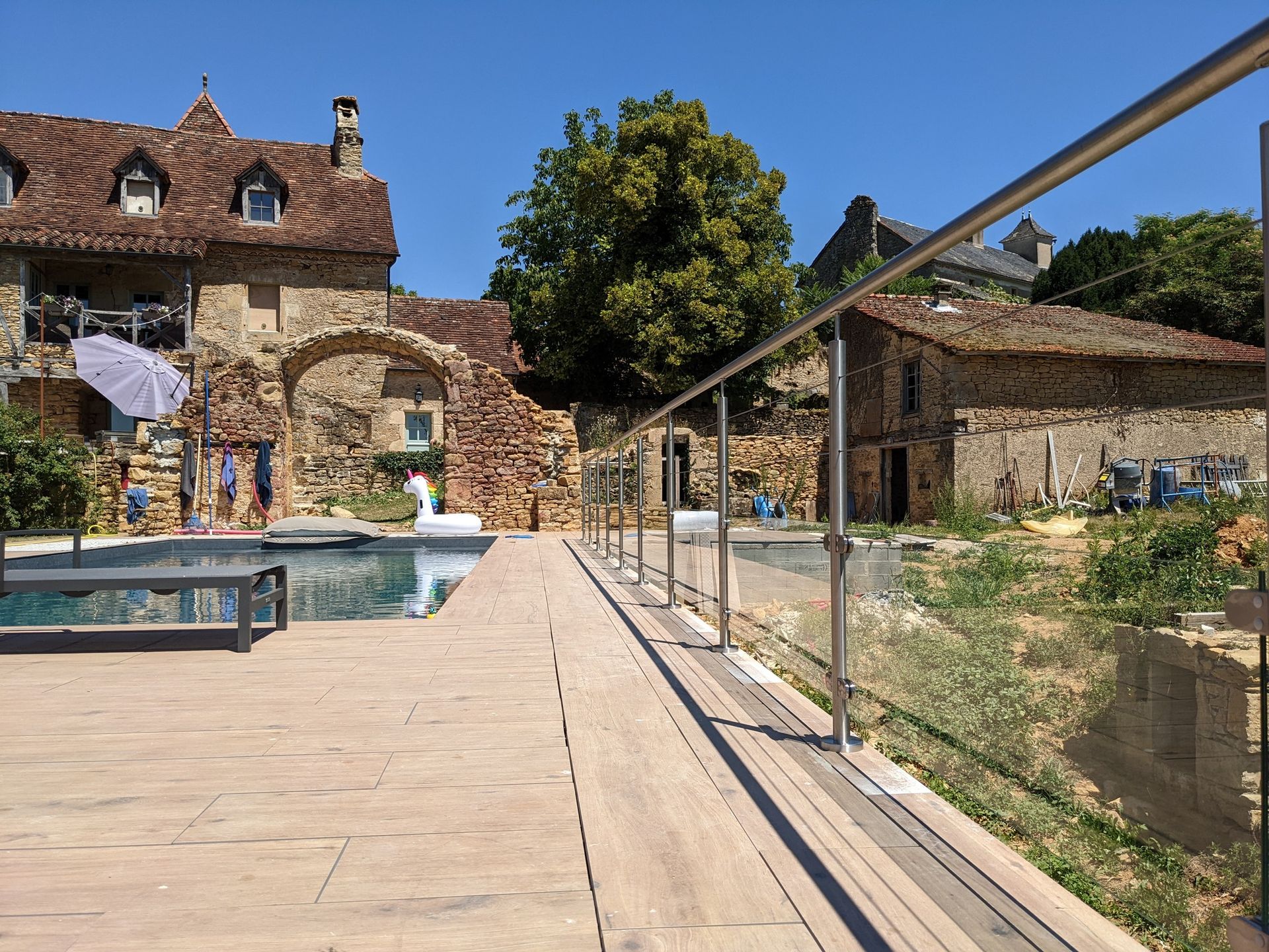 Cour en pierre avec piscine, bâtiments rustiques et passerelle à balustrade de verre sous un ciel bleu azur.