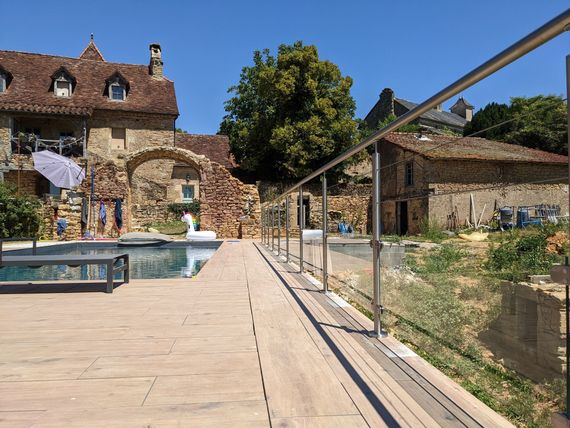 Cour en pierre avec piscine, bâtiments rustiques et passerelle à balustrade de verre sous un ciel bleu azur.