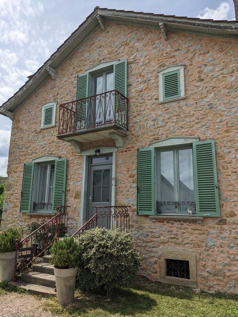 Maison en pierre avec volets verts et balcon, vue de face sous un ciel nuageux