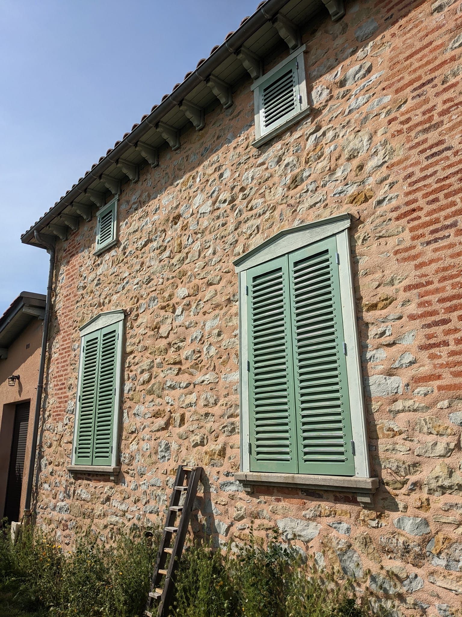 Vieille maison en briques aux volets vert pâle, une échelle appuyée contre le mur sous un ciel bleu.