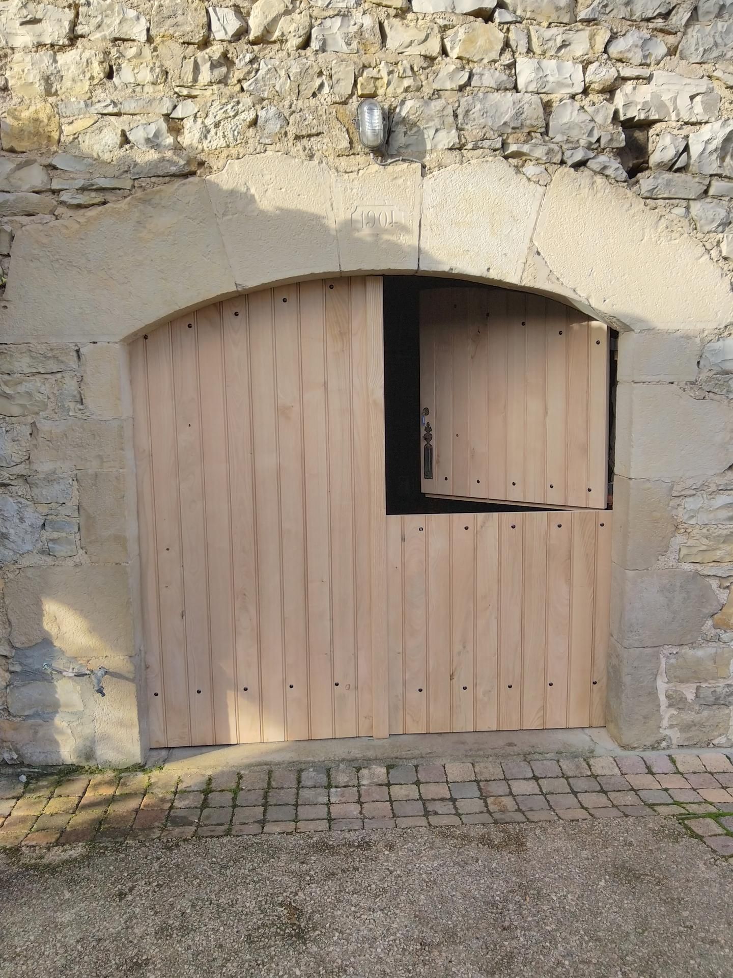 Mur en pierre avec un portail en bois arqué et une petite fenêtre à volets ouverts.
