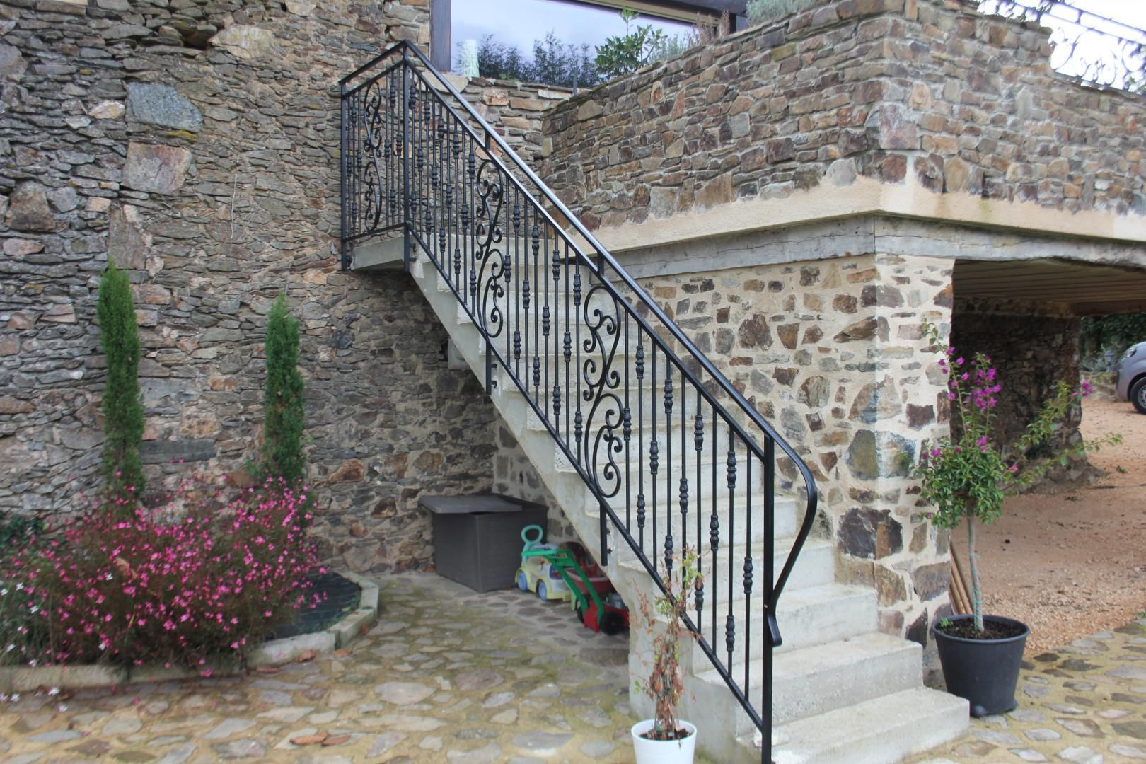 Escalier en pierre avec une rampe noire ornée, jouxtant un bâtiment rustique en pierre et des fleurs en pot.
