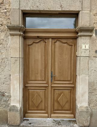 Porte d'entrée double en bois encastrée dans une façade en pierre, surmontée d'une petite imposte.