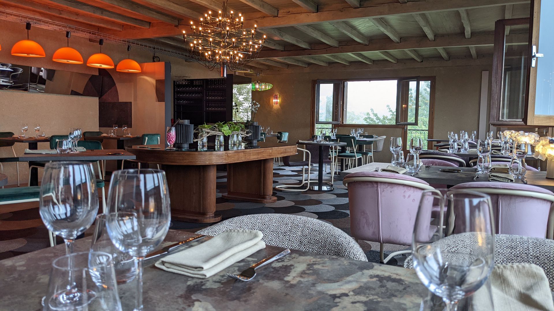 Salle à manger élégante d'un restaurant avec tables dressées, verrerie, lustres et grandes fenêtres donnant sur la verdure.