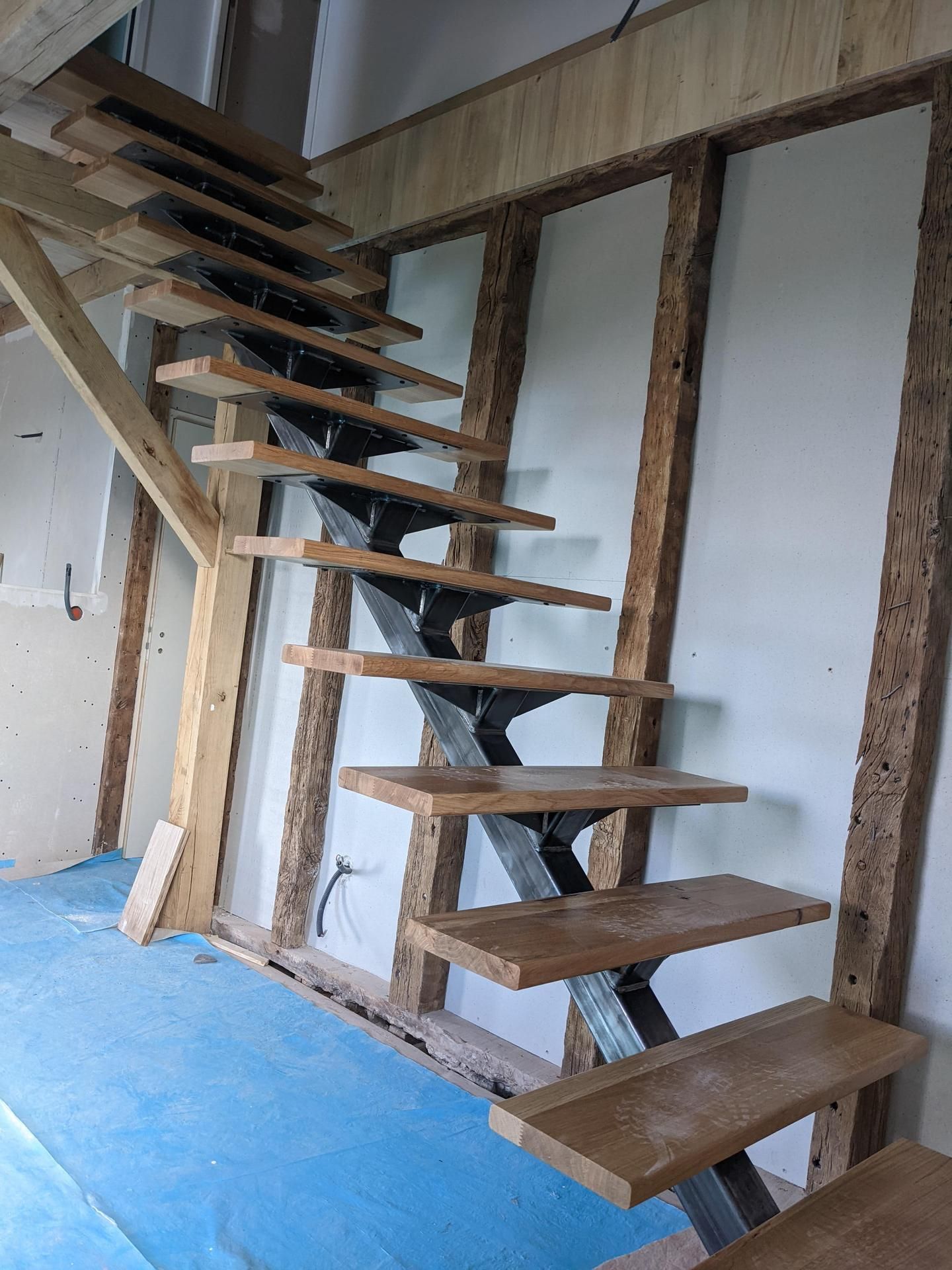 Escalier en bois flottant en construction dans une pièce aux murs à ossature apparente et au sol recouvert de tissu bleu