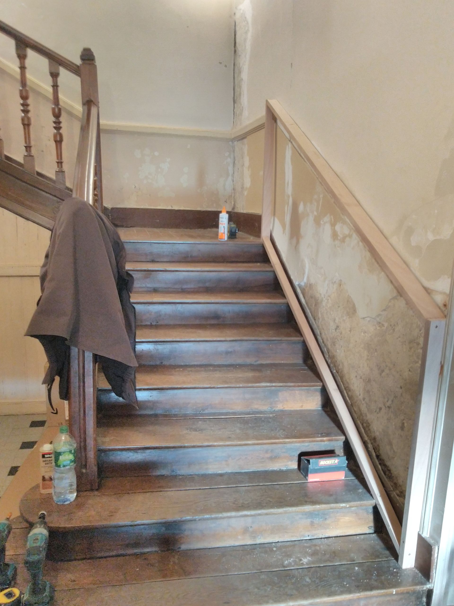 Escalier intérieur inachevé avec murs apparents, un manteau sur la rampe et des objets sur les marches.