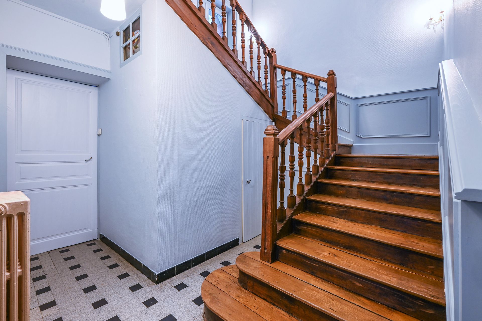 Escalier intérieur avec rampe en bois, porte blanche et sol en carrelage à damier.