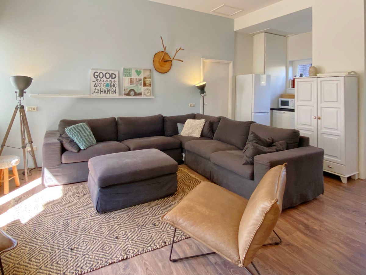 A living room with a grey sectional sofa, matching ottoman, a brown leather armchair, and a kitchen area in the back.