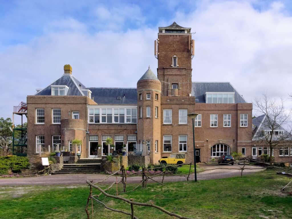 A large, historic brick building with a central tower, multi-level rooflines, and a grassy front area under a blue sky.