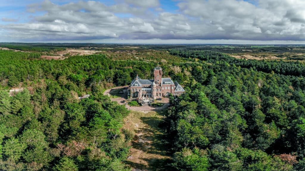 An aerial view of a grand, historic stone castle nestled within a dense, green forest under a cloudy blue sky.