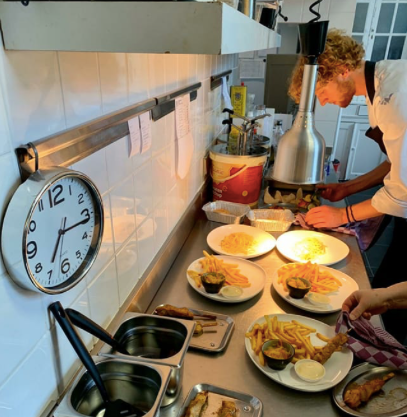 Een keukenmedewerker bereidt borden friet met dipsaus onder een warmtelamp, met een wandklok ernaast.