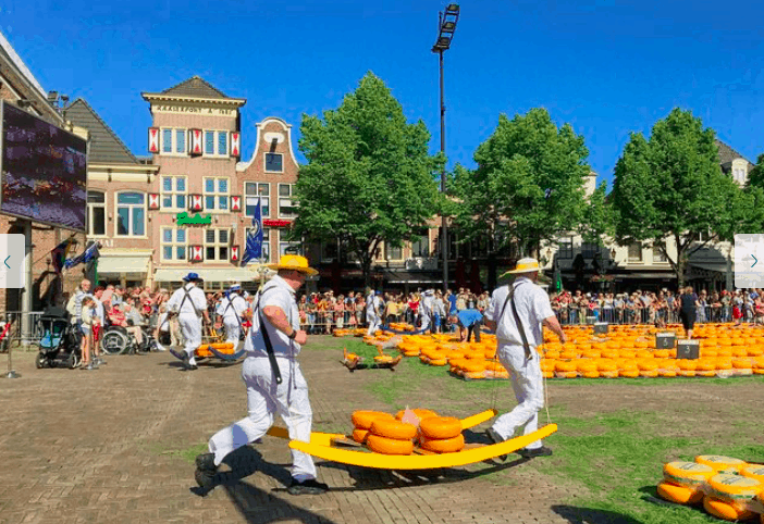 Two people in white uniforms and straw hats carry yellow cheese wheels on a wooden stretcher in a sunny town square.
