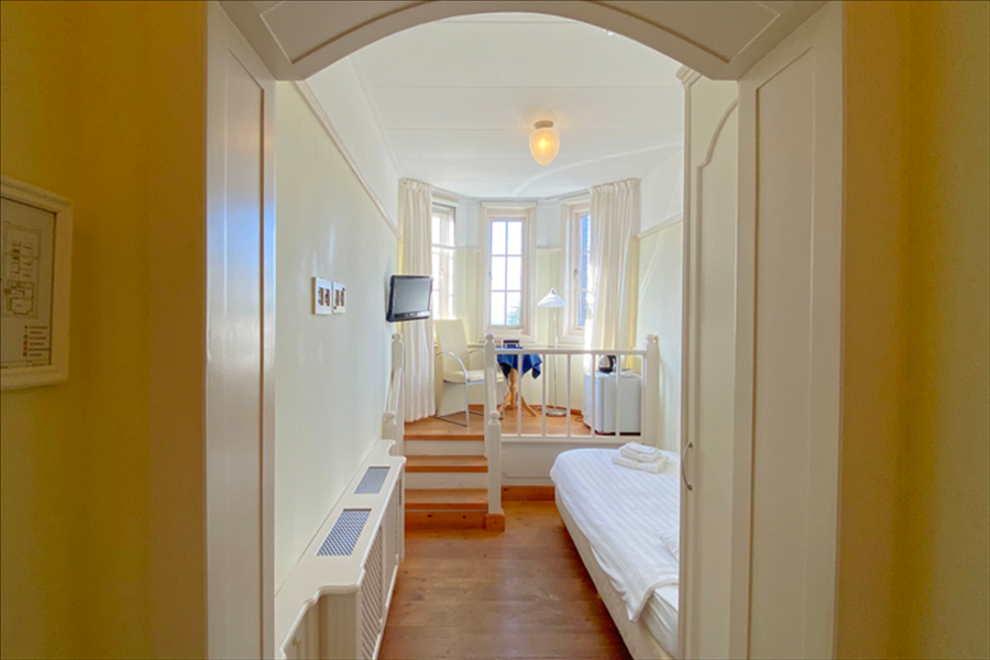 Een hotelkameringang met een gang die leidt naar een verhoogd zitgedeelte bij een erker en een bed aan de rechterkant.