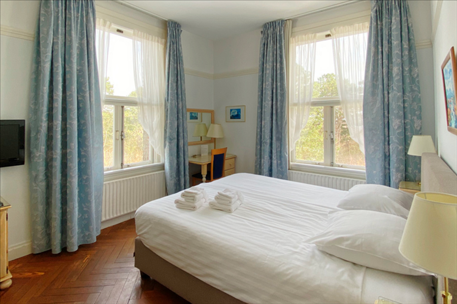 Een slaapkamer met een bed, lichtblauwe gordijnen, houten vloer, twee ramen en een klein bureau in de hoek.