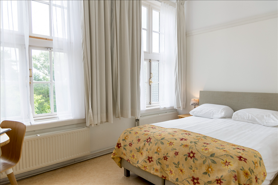 Een lichte slaapkamer met twee grote ramen, witte gordijnen, een bed met een bloemenloper en een houten stoel.