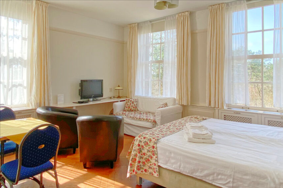 Een hotelkamer met een bed met een bloemenloper, twee donkere fauteuils, een tafeltje, een tv en grote ramen met gordijnen.