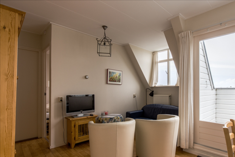 Ein gemütliches Zimmer mit hellen Wänden, zwei weißen Sesseln, einem Fernseher auf einem Holzständer und einer Balkontür mit weißen Vorhängen.