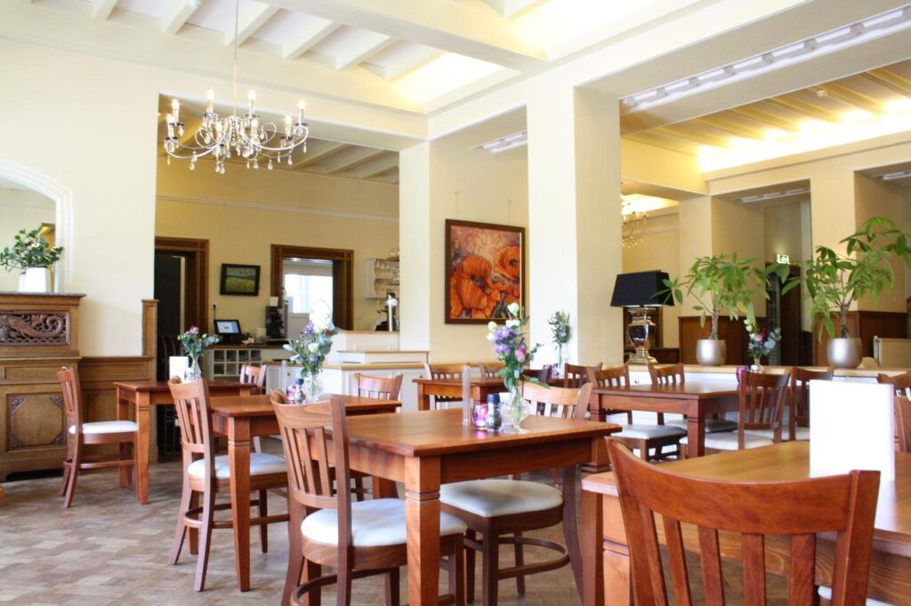 A bright hotel or restaurant dining room with wooden tables, chairs, indoor plants, a chandelier, and wall art.