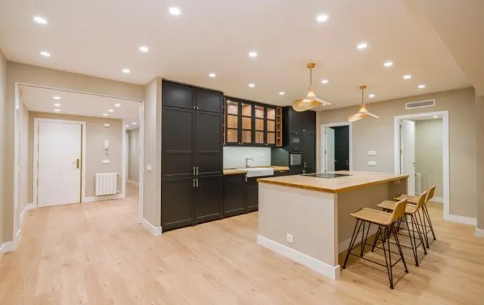 Una cocina moderna con armarios negros, una isla de cocina de madera clara con taburetes y una iluminación cálida empotrada.