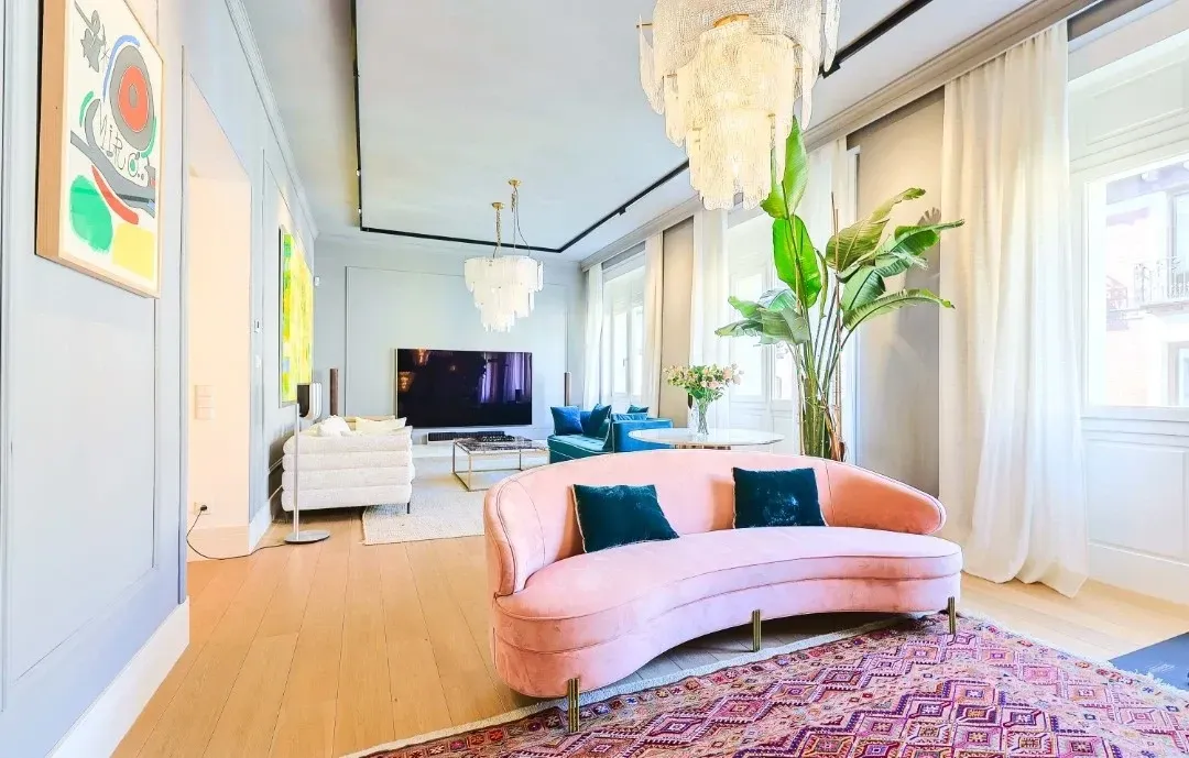 Una sala de estar con un sofá rosa curvo, una alfombra estampada, una planta de interior grande y un televisor al fondo.