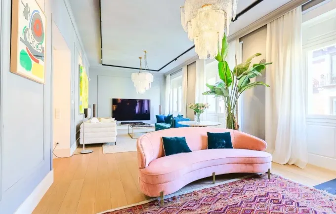 Una sala de estar con un sofá rosa curvo, una alfombra estampada, una planta de interior grande y un televisor al fondo.