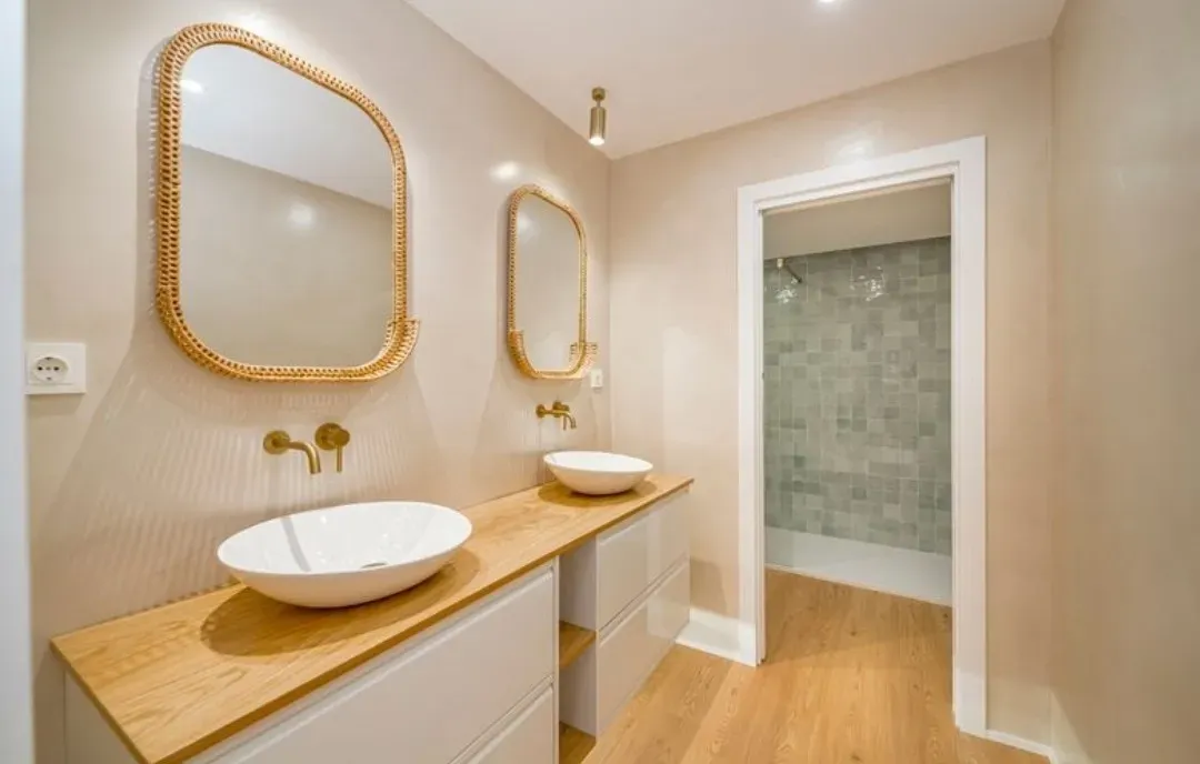 Baño moderno con lavabos dobles sobre encimera, encimera de madera clara, armarios blancos y espejos dorados encima.