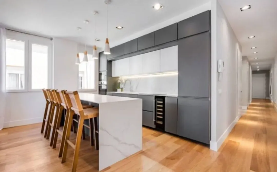 Cocina moderna con isla con encimera de mármol blanco, taburetes de madera, armarios grises y suelos de madera noble.