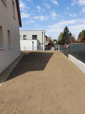 Eine mit Kies befestigte Auffahrt zwischen hellen Gebäuden und einem Zaun unter blauem Himmel.