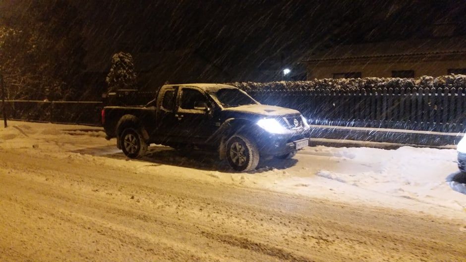 Ein schwarzer Pickup fährt nachts mit eingeschalteten Scheinwerfern auf einer schneebedeckten Straße. Es schneit.
