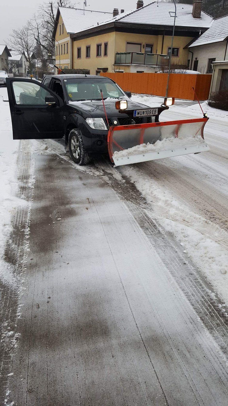 Schwarzes Fahrzeug mit Schneepflug räumt eine schneebedeckte Straße in einer Stadt. Tür geöffnet.