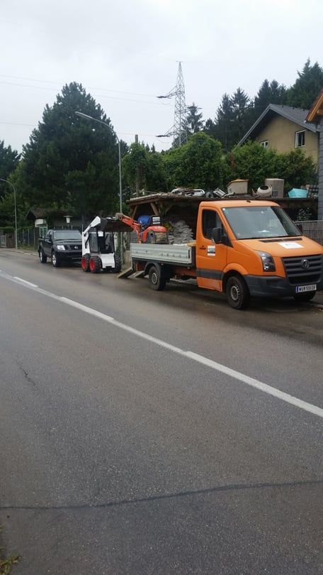 Ein orangefarbener Lastwagen mit Ausrüstung, ein kleiner weißer Traktor und ein schwarzes Fahrzeug parken auf einer Straße.