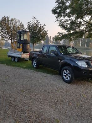 Schwarzer Pickup-Truck zieht einen Anhänger mit einem kleinen Bagger auf einer Schotterstraße an einem See entlang.