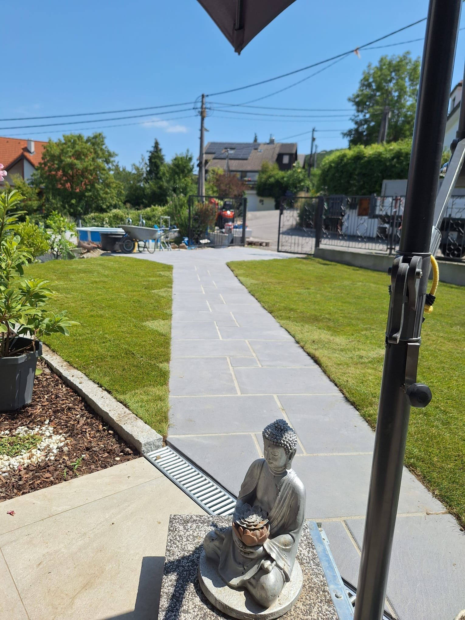 Ein Steinpfad führt durch eine grüne Rasenfläche zu einem Tor; eine kleine Statue steht am Wegesrand. Sonniger Tag.