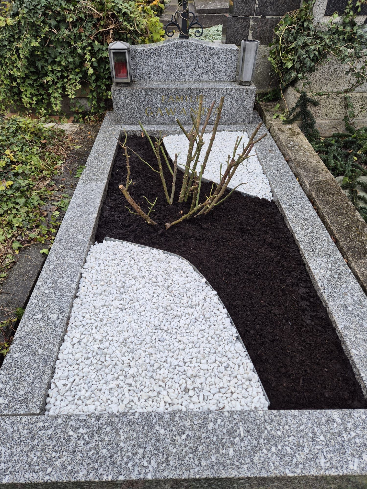 Grab mit Granitgrabstein und -einfassung. Dunkler Mulch und weiße Kieselsteine ​​umgeben einen kahlen Rosenstrauch.