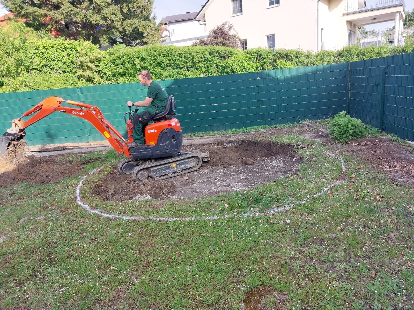 Ein Mann bedient einen kleinen Bagger und hebt eine kreisförmige Fläche in einem grasbewachsenen Hinterhof aus.