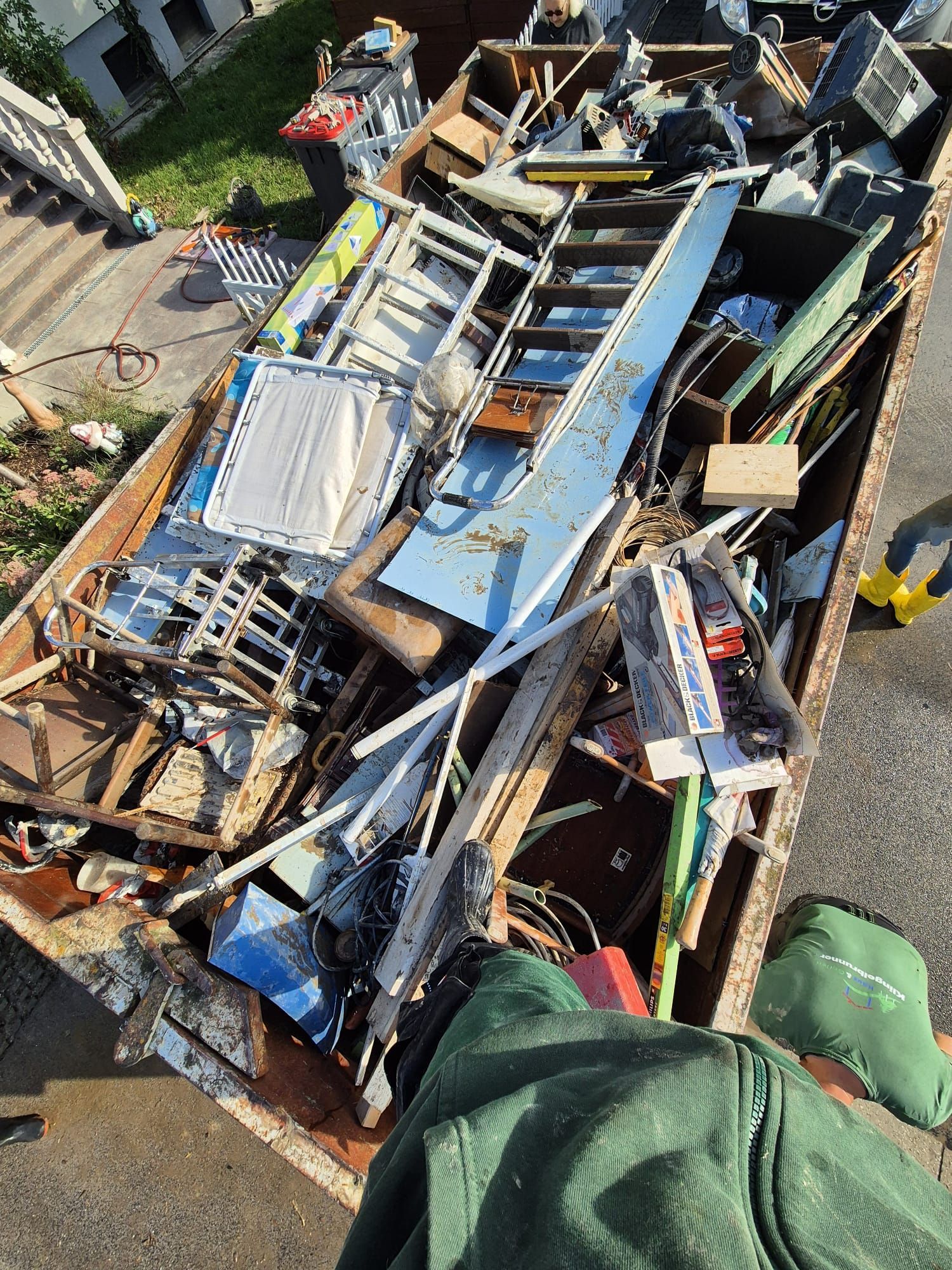 Eine Person steht neben einem Müllcontainer, der überquillt von Bauschutt, darunter eine Leiter, eine blaue Platte und Metallreste.