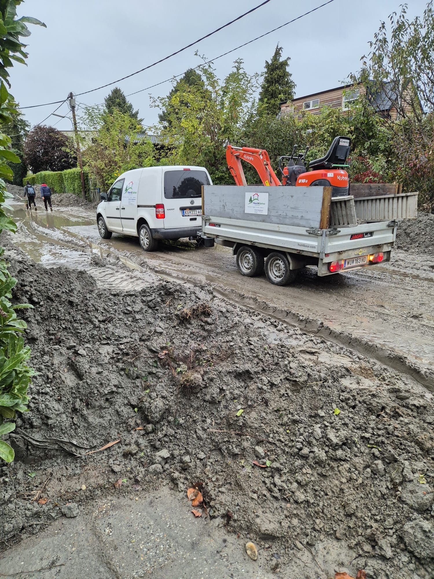 Ein weißer Lieferwagen zieht einen Anhänger mit einem Bagger auf einer matschigen Straße.