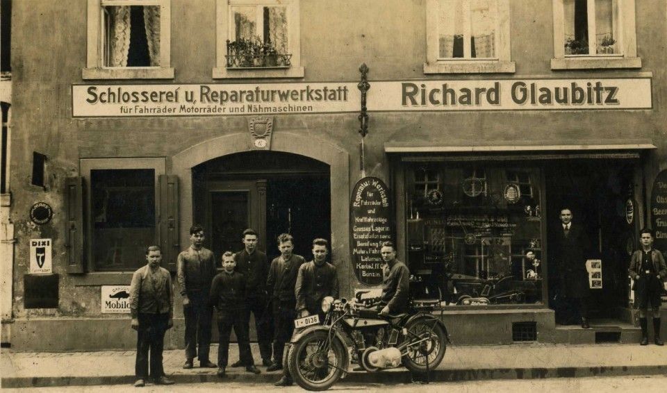Alte Ladenfronten, vor denen sich Menschen versammelt haben, und ein davor geparktes Motorrad mit der Aufschrift „Richard Glaubitz“.