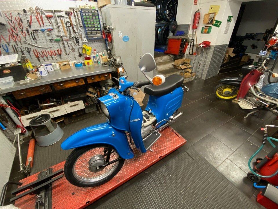 Ein blauer Oldtimer-Roller in einer vollgestellten Werkstatt, in deren Nähe sich Werkzeuge und Motorradteile befinden.