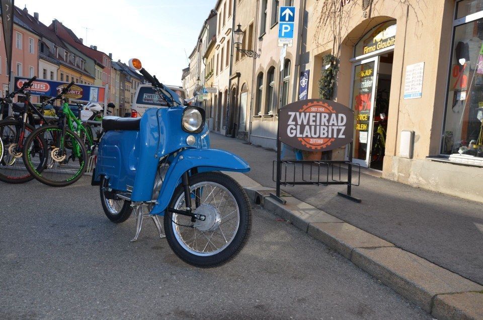 Ein blaues Oldtimer-Motorrad parkt auf einer Straße in der Stadt neben einem Gebäude mit einem Schild mit der Aufschrift „ZWEIRAD GLAUBITZ“.