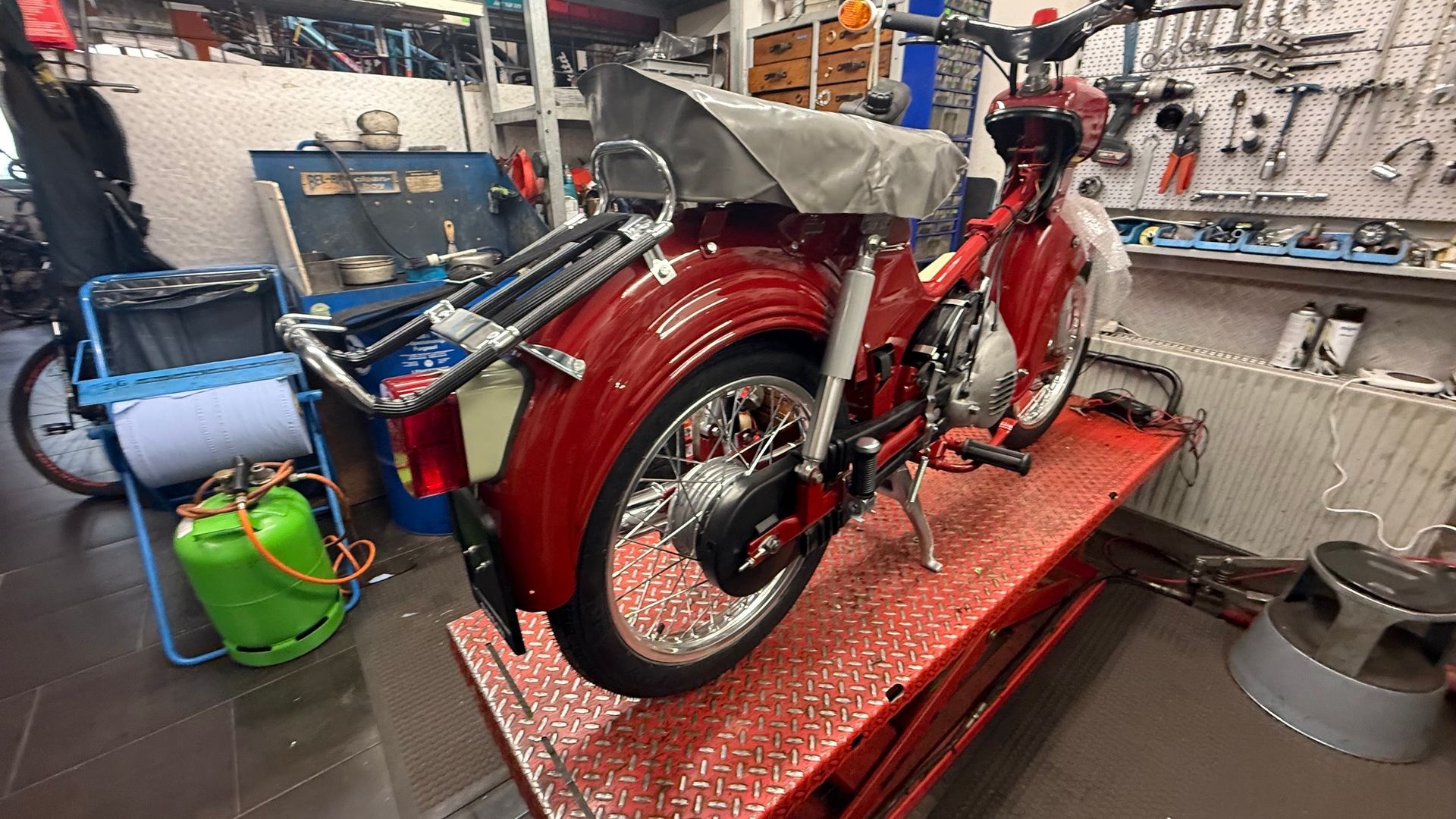 Ein rotes Vintage-Motorrad auf einer Hebebühne in einer Werkstatt, umgeben von Werkzeugen und Ausrüstung in einer Reparaturwerkstatt