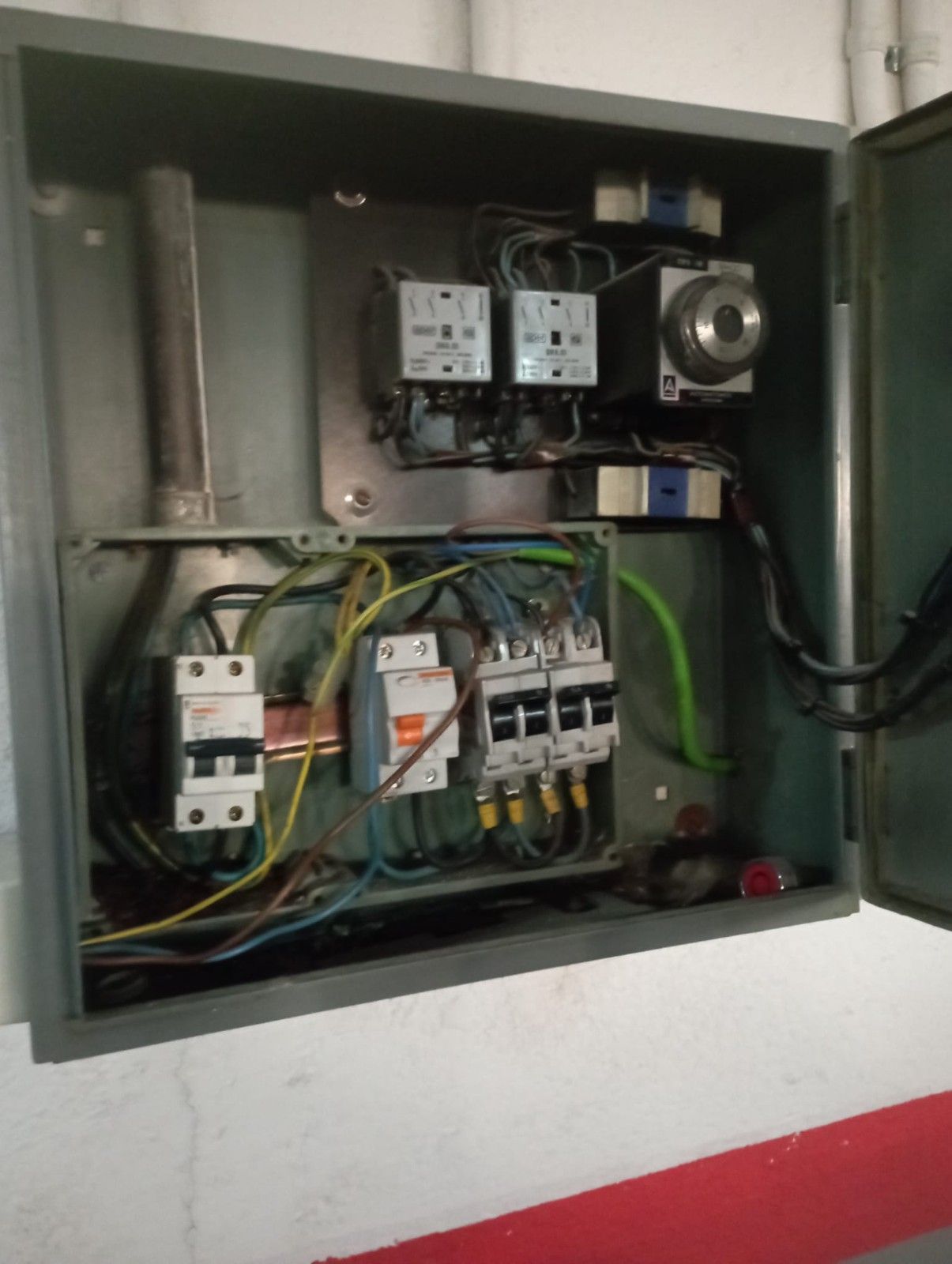 Caja de control eléctrico gris abierta en una pared que contiene múltiples disyuntores, relés y cableado interno expuesto.