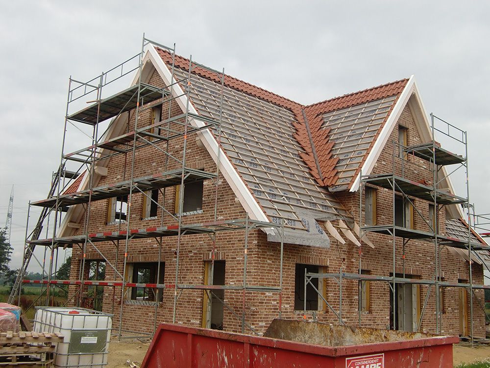 Es wird ein Backsteinhaus mit Gerüst gebaut, davor steht ein roter Müllcontainer.