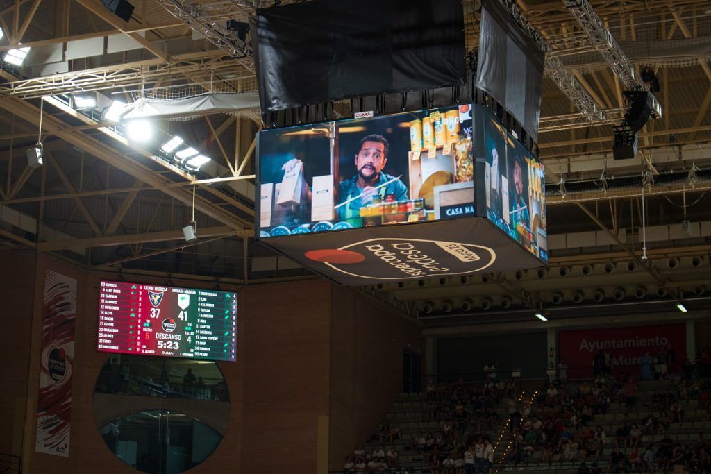 Un gran estadio cubierto con múltiples pantallas que muestran contenido. La gente observa desde abajo mientras se muestran imágenes y gráficos.