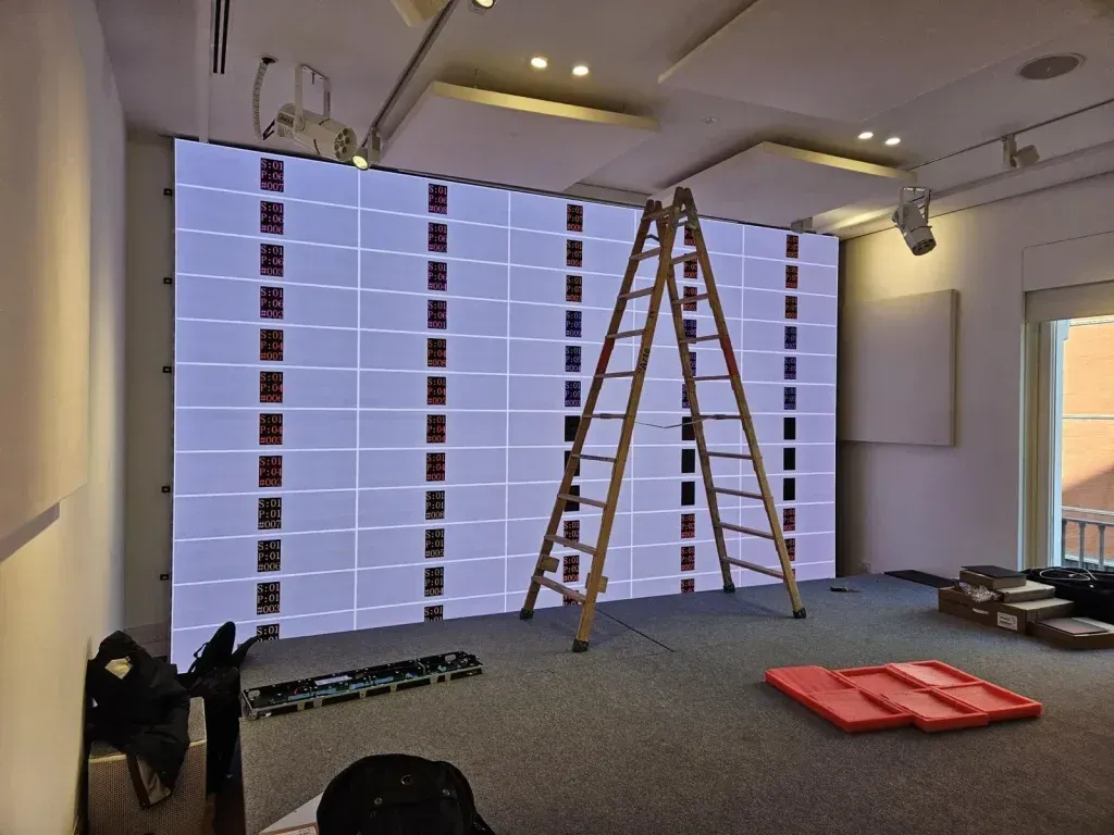 Una escalera de madera se apoya contra una gran pantalla iluminada en una habitación. Se ven patrones rojos y blancos en la pantalla.