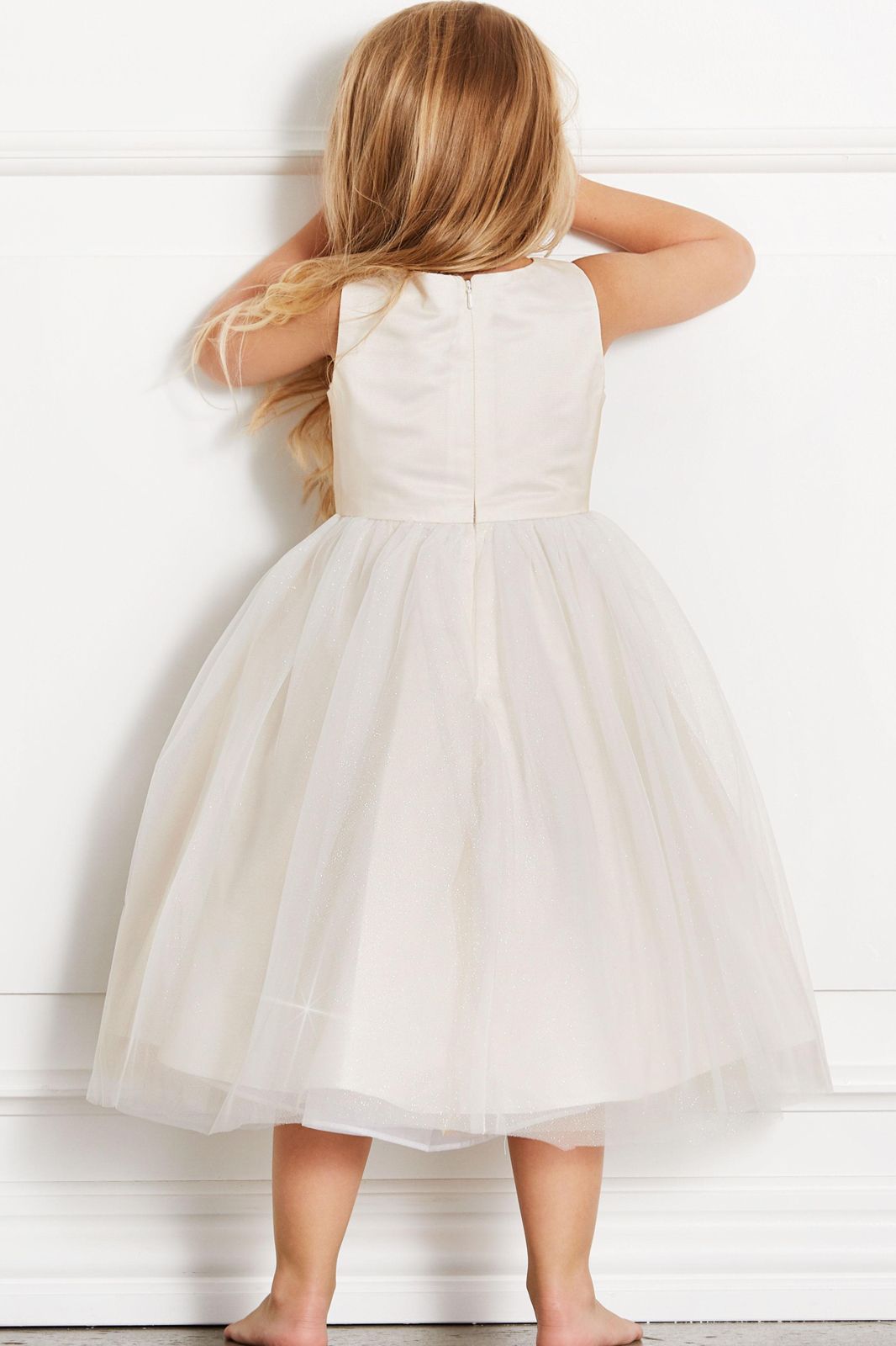 Ein kleines Mädchen in einem weißen Kleid steht vor einer weißen Wand.