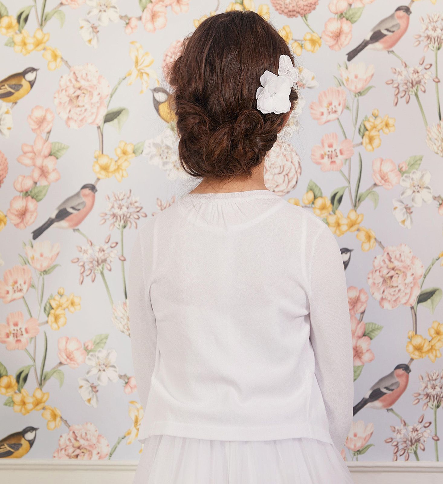 Eine Frau in einem weißen Kleid steht vor einer Wand mit Blumen und Vögeln darauf.